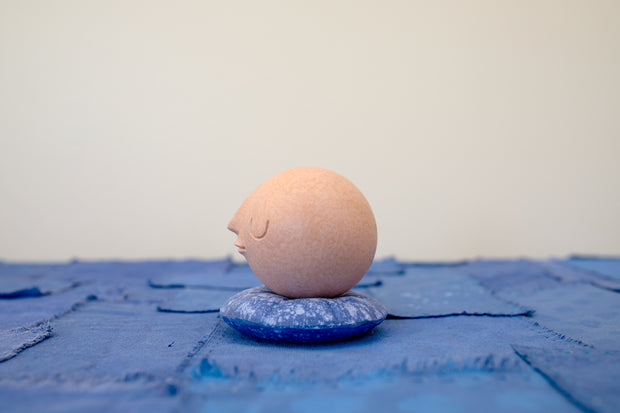 Small carved wooden sculpture of a tannish orange sphere with a closed eye, calm smiling expression. It rests atop of a rounded purple wooden pillow with splatter patterns, carved to look soft.