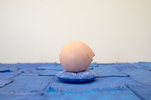 Small carved wooden sculpture of a tannish orange sphere with a closed eye, calm smiling expression. It rests atop of a rounded purple wooden pillow with splatter patterns, carved to look soft.