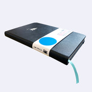 Side view of black pleather notebook with a paper sleeve wrapped around it and a blue ribbon as a bookmark.