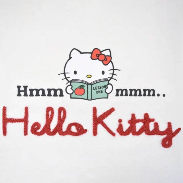 White long sleeve shirt with a graphic in the center of Hello Kitty reading a book. Around her says "Hmmmmm.." with half the text on one side and the other half on the other. Below is a red yarn embroidered element spelling out "Hello Kitty".