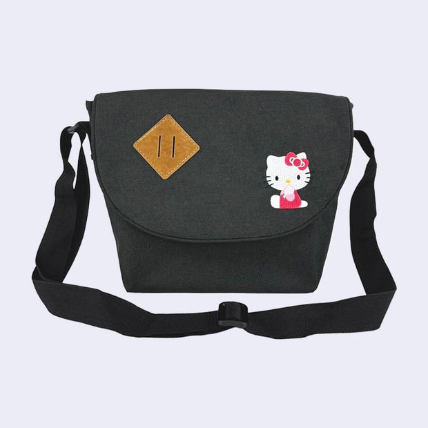 Dark gray messenger bag with an embroidery of Hello Kitty, sitting on the ground and looking to the side with her hand over her mouth. The back has an extendable strap handle and a brown leather off tilt square patch.