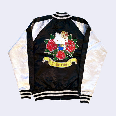 Back view of Sukajan style bomber jacket with a black semi shiny body with ivory colored sleeves and color accents. Bottom of the sleeves and jacket have a white and black striped band. A large graphic of Hello Kitty with red roses is on the back.