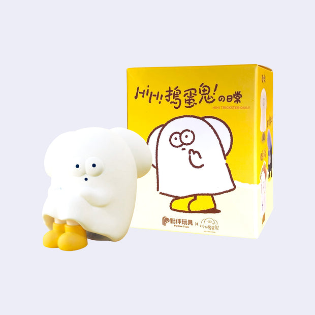 Small vinyl figure of a white cartoon ghost with floppy bunny like ears, yellow boots and a surprised expression. It sits with its knees pulled in, in front of its product packaging.