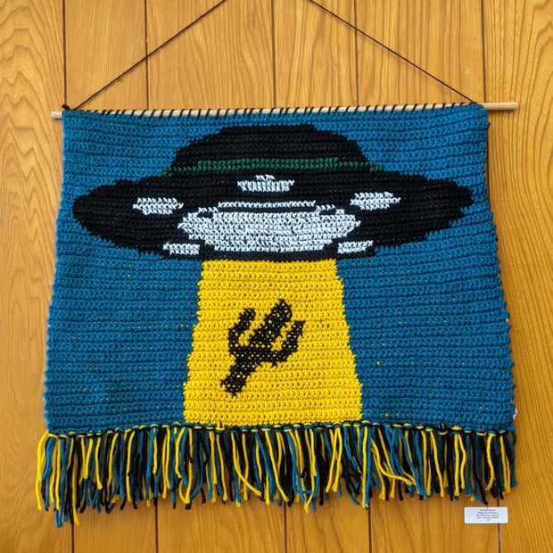 Wall hanging crochet tapestry of a large UFO abducting a cactus.