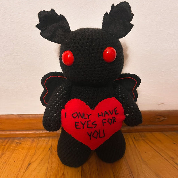 Sculpture of a black cartoon style moth with large red bead eyes and rounded limbs. It holds a red heart that reads "I only have eyes for you"