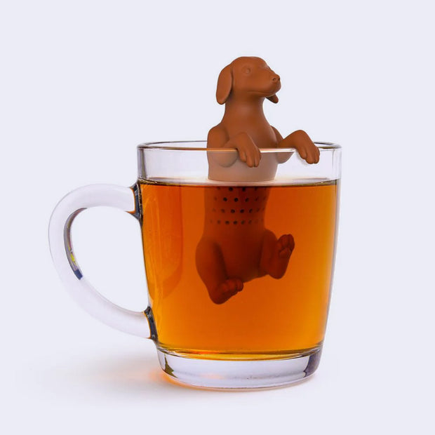 Brown silicone dachshund dog sitting in a cup of tea with holes in its body to diffuse the tea leaves. Its front paws hang over the edge of the glass.