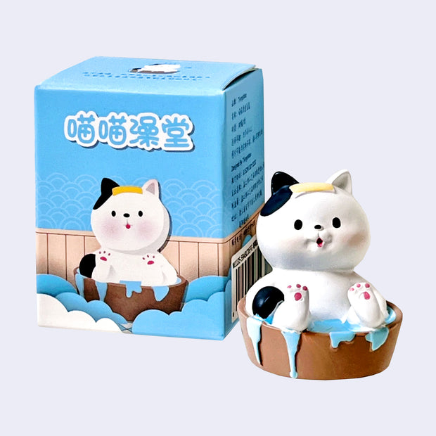 Figurine of a white cat with black spots in a cute, cartoon style. It soaks in a small wooden bath with a yellow cloth atop its head. It sits next to its product packaging.