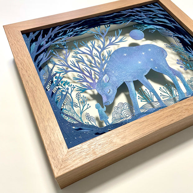 Paper cutting diorama of a large blue deer, their antlers having many, many separate branches. A small girl stands and touches the nose of the bowing deer. They are surrounded by trees and cut paper plants.