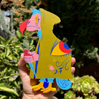 Die cut wooden sculpture of a Dodo bird with an olive green hooded cloak and an eye on its back. It holds a dagger, faced downwards.