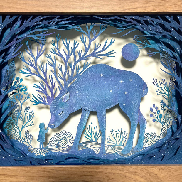 Paper cutting diorama of a large blue deer, their antlers having many, many separate branches. A small girl stands and touches the nose of the bowing deer. They are surrounded by trees and cut paper plants.