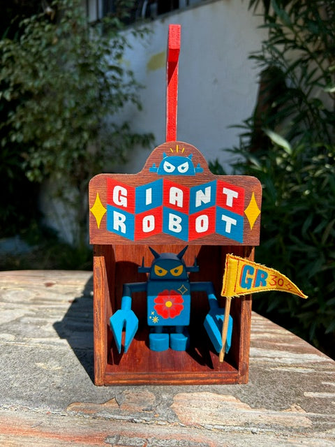 Painted wooden showcase box that holds a blue wooden robot inside. Exterior reads "Giant Robot" written in stylistic retro font, red blue and white. Wooden robot has a flower on its stomach and holds a yellow flag.