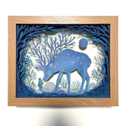 Paper cutting diorama of a large blue deer, their antlers having many, many separate branches. A small girl stands and touches the nose of the bowing deer. They are surrounded by trees and cut paper plants.