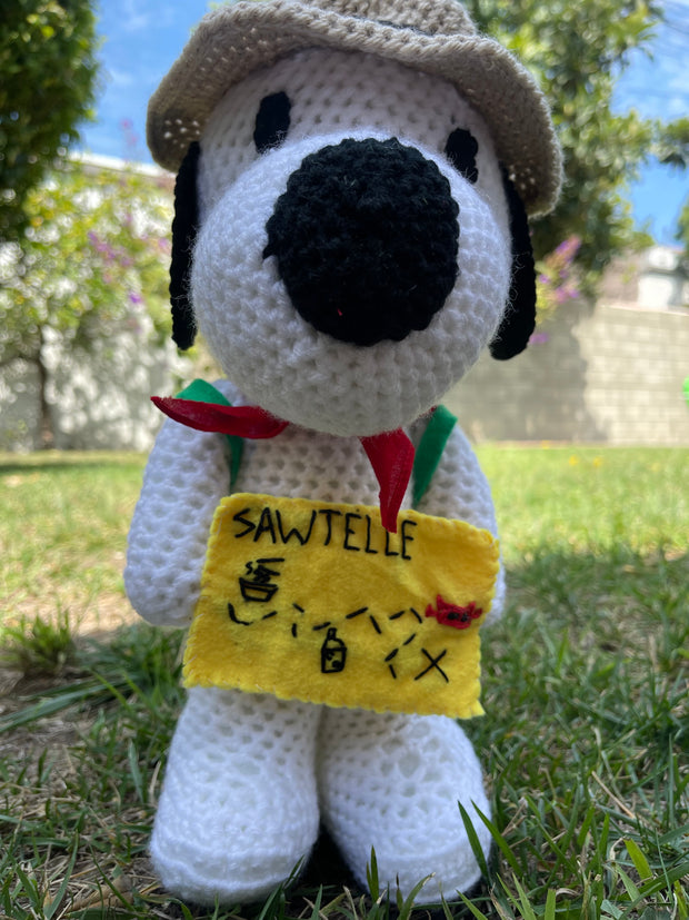 Crocheted plush doll of Snoopy with a brown outdoors hat, a red scarf and a green backpack. He holds a yellow felt map of Sawtelle.