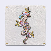 Enamel pin with gold outlining of a pastel colored snake, with its body elongated with small twists. The pattern of its scales is floral in nature and 3 flowers with blooming from the snake's body. Pin is attached to an elaborately designed all white backing card.