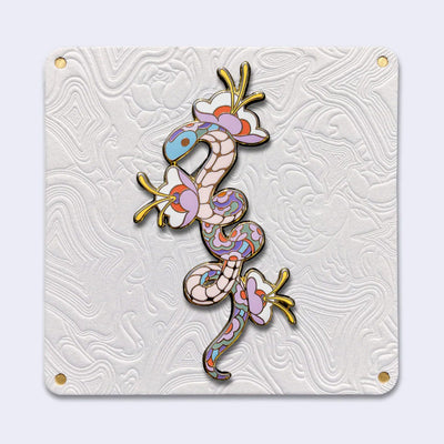 Enamel pin with gold outlining of a pastel colored snake, with its body elongated with small twists. The pattern of its scales is floral in nature and 3 flowers with blooming from the snake's body. Pin is attached to an elaborately designed all white backing card.