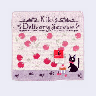 Square towel, cream and light pink striped with cherries. Top has stylized text reading "Kiki's Delivery Service" and bottom has paw prints, a cupcake and Jiji the black cat.