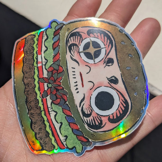 Die cut sticker of a burger shaped to look like a Japanese daruma doll with a gold and red braided rope around the center. Shown at an angle to display holographic sheen.