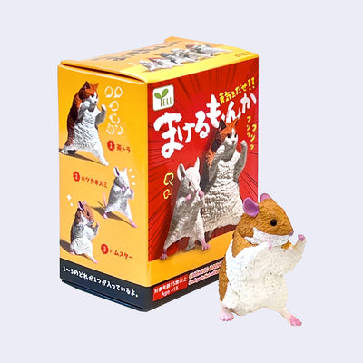 Small plastic figure of a hamster, in a fighting stance with its fists up. It stands in front of its product packaging.