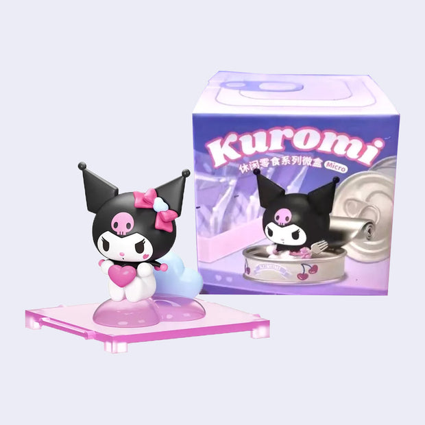 Figure of Kuromi, holding a heart and sitting atop of a clear pink heart. It sits in front of its product packaging.