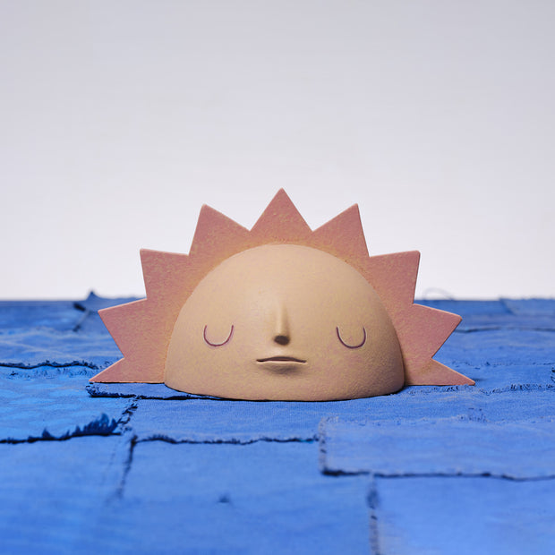 Carved wooden sculpture of a rising sun, visible with only its top half showing. It is a muted yellow and orange with pointed sun rays around it and a calm, closed eye facial expression.