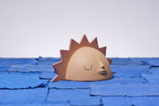 Carved wooden sculpture of a rising sun, visible with only its top half showing. It is a muted yellow and orange with pointed sun rays around it and a calm, closed eye facial expression