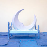 Carved wooden sculpture of a large, sharp tipped purple crescent moon with a closed eye calm expression. It rests atop of a blue bed on a dark blue wooden bed frame. Purple stars rest on the bed as well.