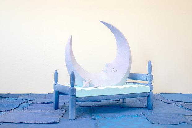 Carved wooden sculpture of a large, sharp tipped purple crescent moon with a closed eye calm expression. It rests atop of a blue bed on a dark blue wooden bed frame. Purple stars rest on the bed as well.