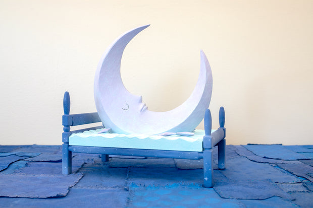 Carved wooden sculpture of a large, sharp tipped purple crescent moon with a closed eye calm expression. It rests atop of a blue bed on a dark blue wooden bed frame. Purple stars rest on the bed as well.