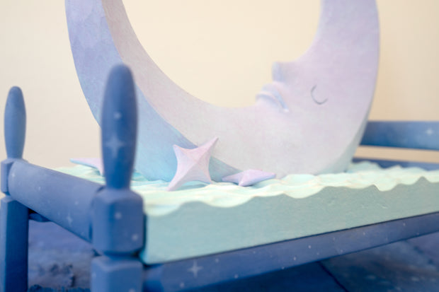 Carved wooden sculpture of a large, sharp tipped purple crescent moon with a closed eye calm expression. It rests atop of a blue bed on a dark blue wooden bed frame. Purple stars rest on the bed as well.