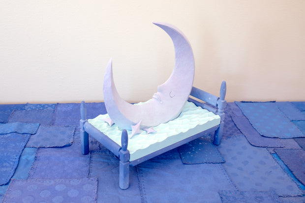 Carved wooden sculpture of a large, sharp tipped purple crescent moon with a closed eye calm expression. It rests atop of a blue bed on a dark blue wooden bed frame. Purple stars rest on the bed as well.