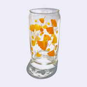Glass cup with a wrap around graphic of orange and yellow gingko leaves and thin purple flowers. A thin, nude woman without many facial or body features sits in the center atop a leaf.