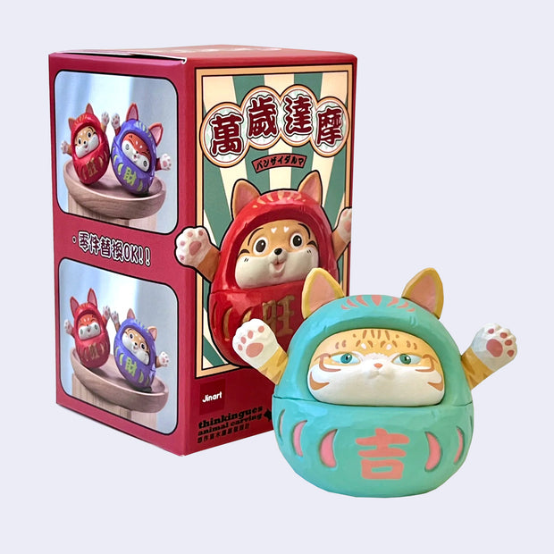 Small figure of a orange tabby cat in a daruma doll costume, light teal with pink color accents. It has its arms raised up and a calm expression. It stands next to its product packaging.