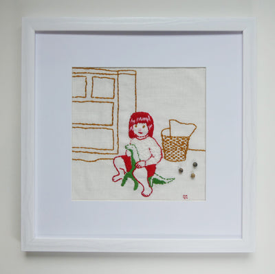 Framed embroidery of a small girl with red hair and red outline, wearing a white long sleeves and red shorts. She sits on the ground of a bedroom and plays with a green dinosaur doll.