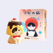 Figure made to look like its been carved, of a cute calico cat with a red bowtie and a pudding dessert around its head like a hood hat. It sits and smiles, next to its product packaging.