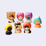 7 small figures of cats, designed to look like they're carved from wood and painted, all sitting and smiling with objects around their heads like hood hats. Hats are: bread, pink tulip, purple tulip, mountain, lemon, pudding and apple.