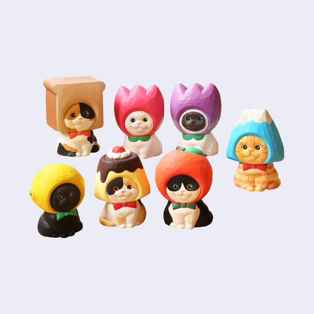 7 small figures of cats, designed to look like they're carved from wood and painted, all sitting and smiling with objects around their heads like hood hats. Hats are: bread, pink tulip, purple tulip, mountain, lemon, pudding and apple.