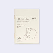 Cream colored blank cover journal within packaging with a cream insert that has Japanese writing and an illustration of a hand holding 3 notebooks.