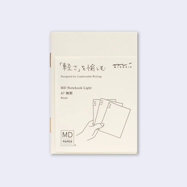 Cream colored blank cover journal within packaging with a cream insert that has Japanese writing and an illustration of a hand holding 3 notebooks.