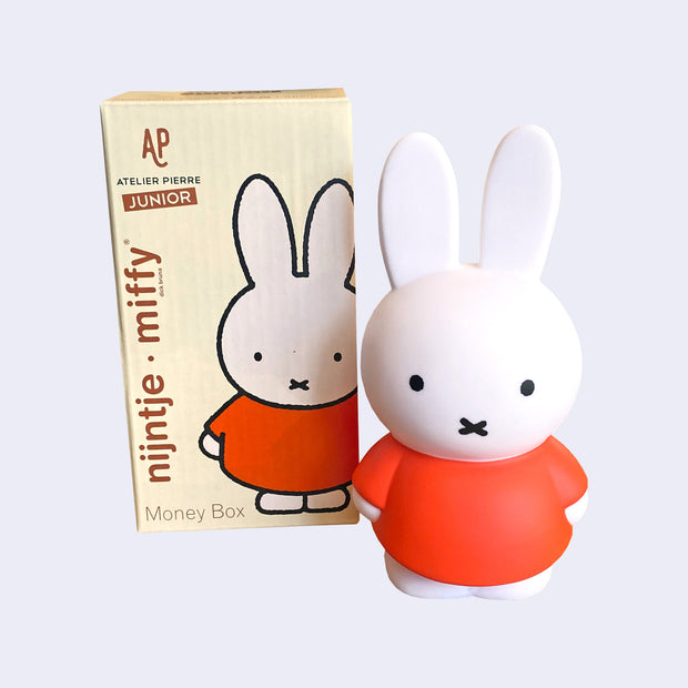 Coin bank shaped like Miffy, wearing her standard reddish orange dress. She stands next to the product packaging.