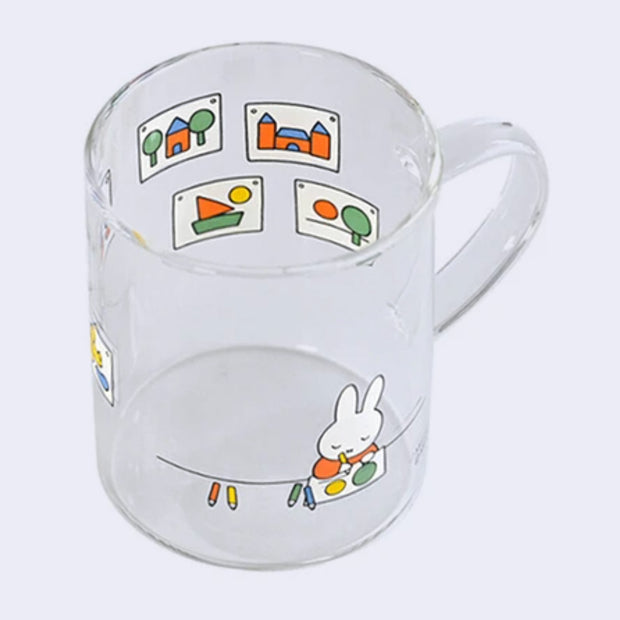 Glass mug with a wrap around graphic of Miffy, sitting and drawing with colored pencils. All around are hung up drawings of cute, simple subject matter.