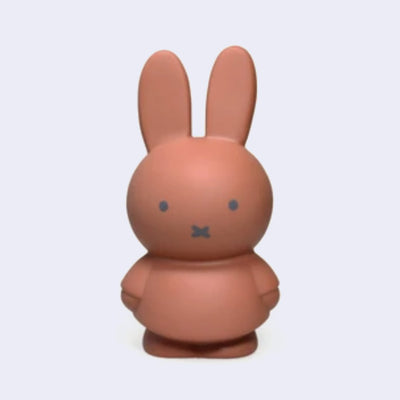 Miffy shaped coin bank, completely colored a terracotta orangish-brown with gray eyes and mouth. She stands wearing a dress with her arms at her sides.