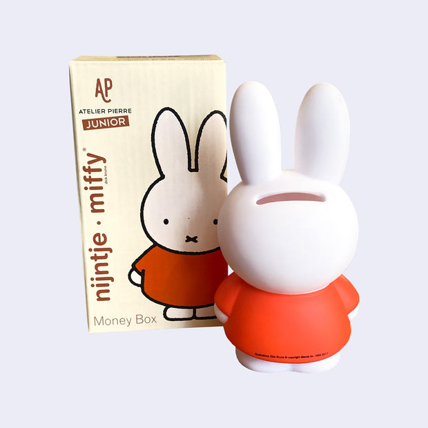 Coin bank shaped like Miffy, wearing her standard reddish orange dress, with a slit in the back of the head for coins. She stands next to the product packaging.