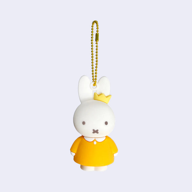 Rubber mascot figure of Miffy, wearing a collared orange dress and a small crown over one of her ears. The figure is attached to a gold toned ball chain.