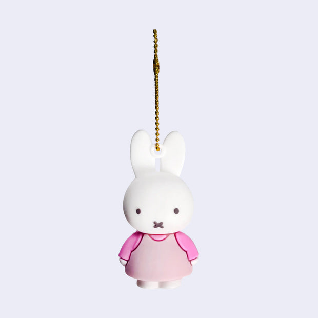 Rubber mascot figure of Miffy, wearing a light pink dress with a pink long sleeve underneath. The figure is attached to a gold toned ball chain.