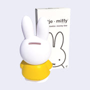 Coin bank shaped like Miffy, wearing her standard yellow dress, the back of her head has a coin slot. She stands next to the product packaging.