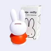 Coin bank shaped like Miffy, wearing her standard orange dress, the back of her head has a coin slot. She stands next to the product packaging.