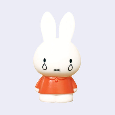 Figure of Miffy, standing with her arms at her side while wearing an orange dress. 2 tears fall out of her eyes as she looks straight forward.