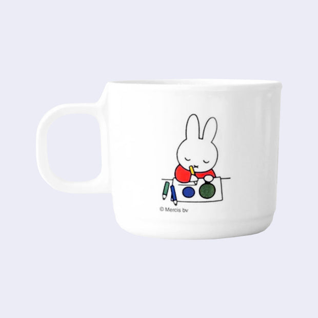 White melamine mug with a graphic of Miffy sitting at a desk and drawing circles on a piece of paper. Her eyes are closed.