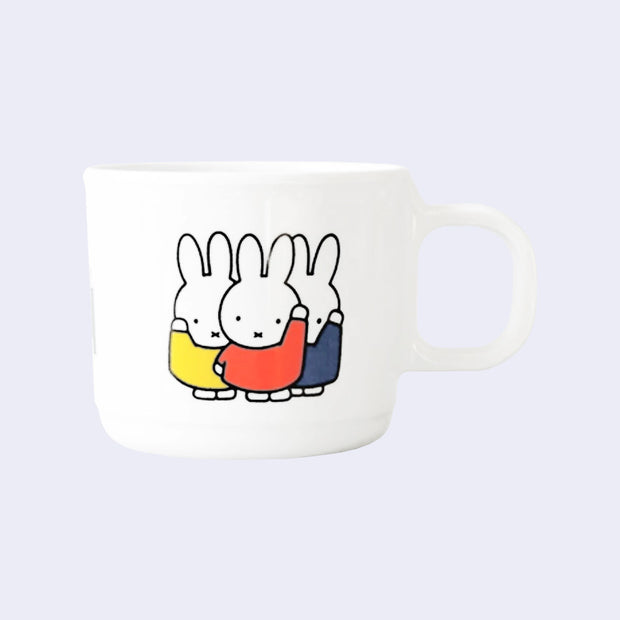 White melamine mug with a graphic of 3 Miffy characters, standing so close they slightly overlap. Each wears a different color shirt - yellow, orange and blue. 