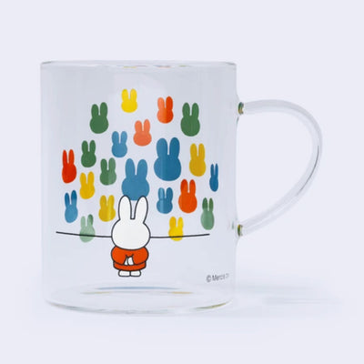 Glass mug with a wrap around graphic of Miffy, standing with her arms behind her back and looking at a large colorful array of solid colored bunny heads on a wall, as though in a museum.